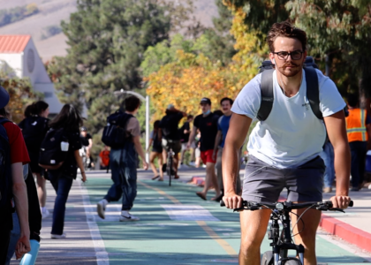 A man rides a bike in a bike lane through Cal Poly's campus. Pedestrians stroll in the background.