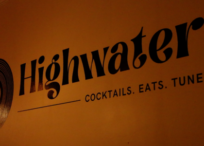 A wall is pictured with the word “Highwater” is a bold font. Under this title are the words “Cocktails. Eats. Tunes.” in all capital letters. A drawing of a record is seen on the left of the text.